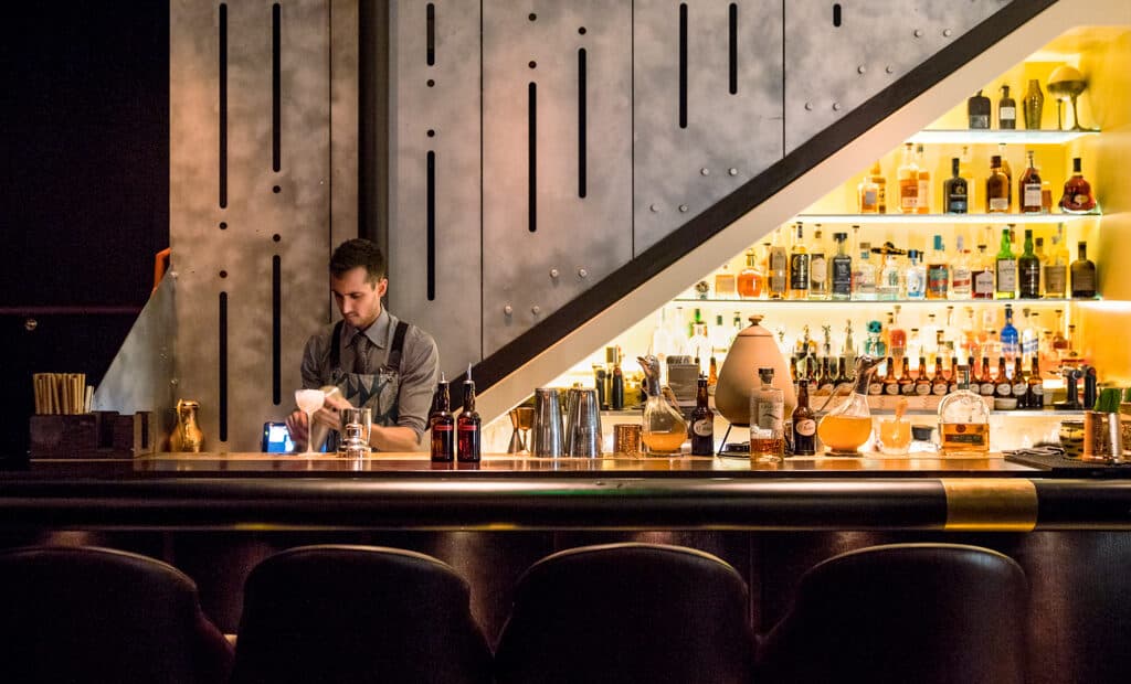 Bartender behind a backlit bar with shelves of bottles