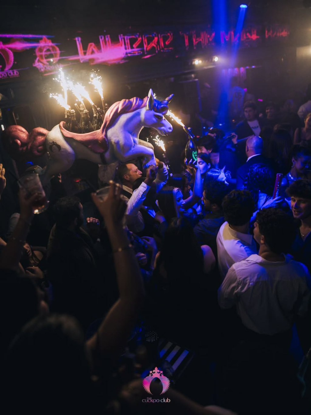 Cuckoo Nightclub table clients dancing while waiters carry a bottle-filled unicorn-shaped bottle presenter through the crowd.